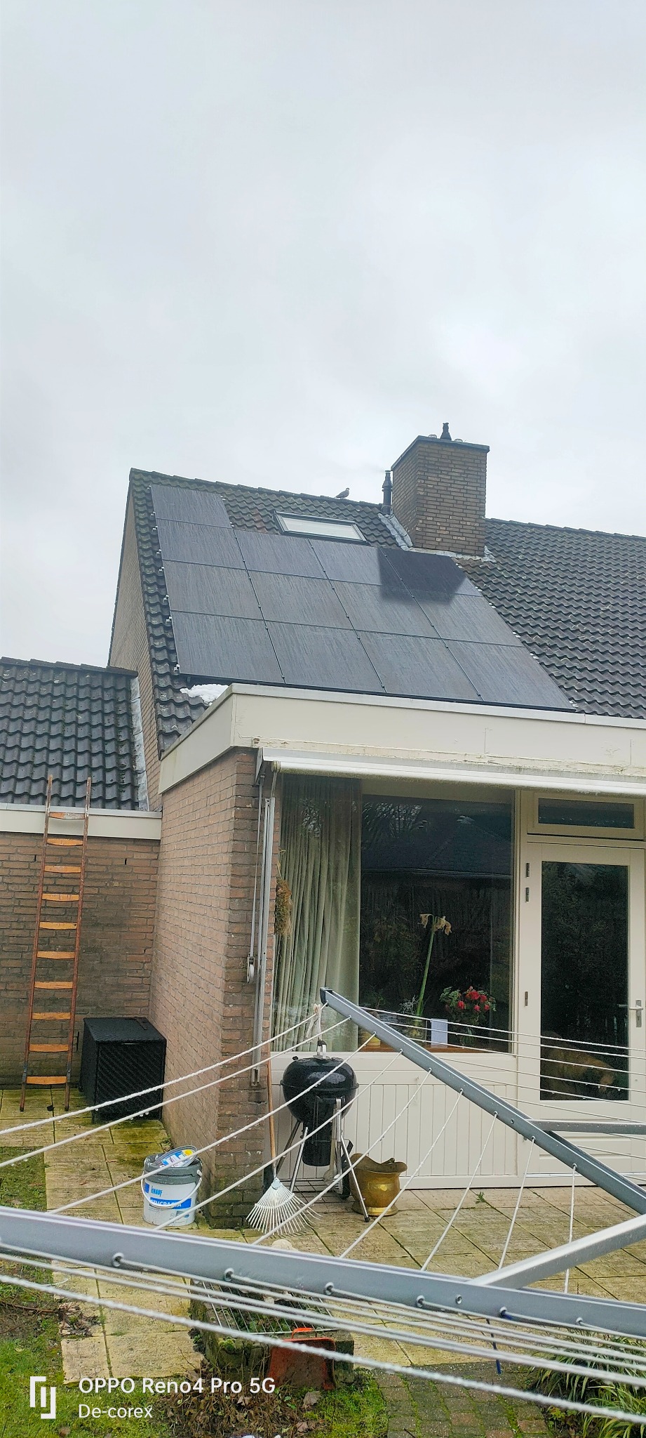 Ciemne panele słoneczne na dachu domu jednorodzinnego w pochmurny dzień, widoczne drabina oparta o ścianę, suszarka na pranie i elementy wyposażenia ogrodu.