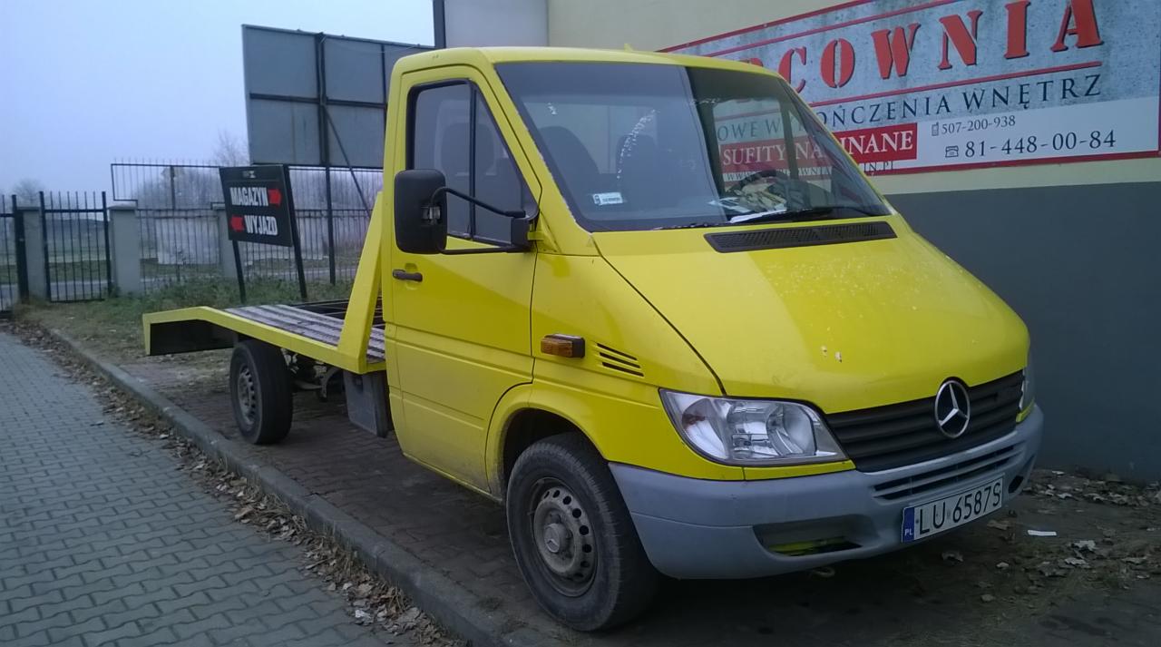 Żółta laweta Mercedes Sprinter z tablicą LU 65879 stoi na chodniku, na tle budynku z reklamą. Widoczny znak 'Magazyn Wyjazd'.