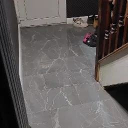 Szare płytki podłogowe imitujące marmur, ułożone w korytarzu przy białych drzwiach wejściowych i drewnianych schodach, z widocznymi butami.