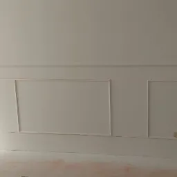 Ściana w skosie z zamontowanymi białymi panelami dekoracyjnymi w formie prostokątnych ramek, widoczny fragment podłogi z drobnymi zabrudzeniami.