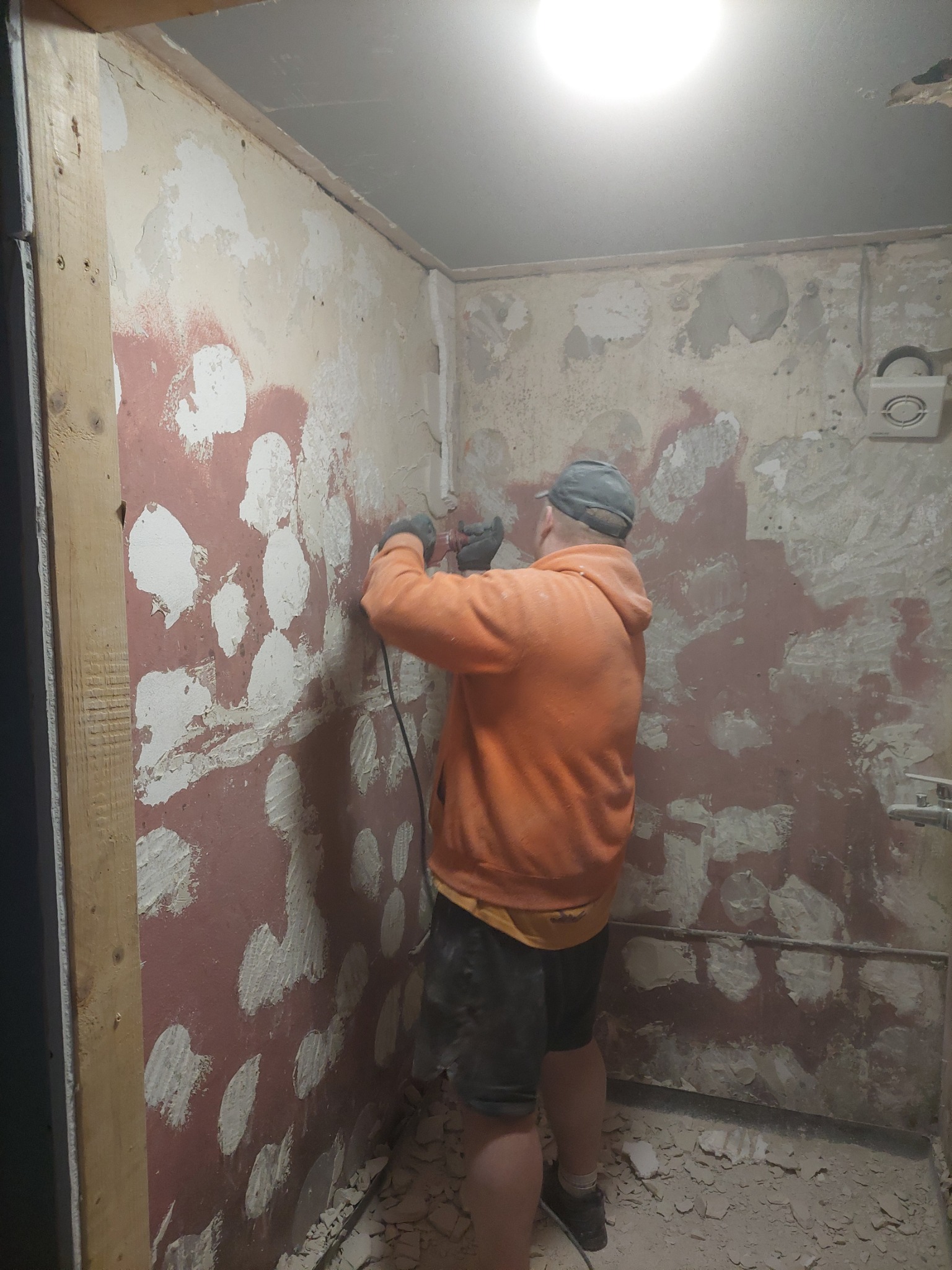 Pracownik w pomarańczowej bluzie szlifuje ścianę w łazience przed położeniem płytek, widoczne ślady po kuciu i pozostałości starej glazury na ścianach i podłodze.