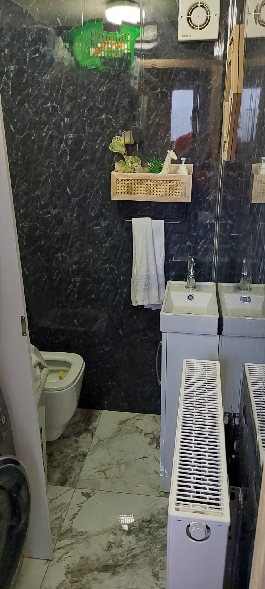 Umywalki nablatowe z szafkami w łazience z czarnymi marmurkowymi płytkami, dekoracyjne akcesoria na półce rattanowej i grzejniki panelowe.