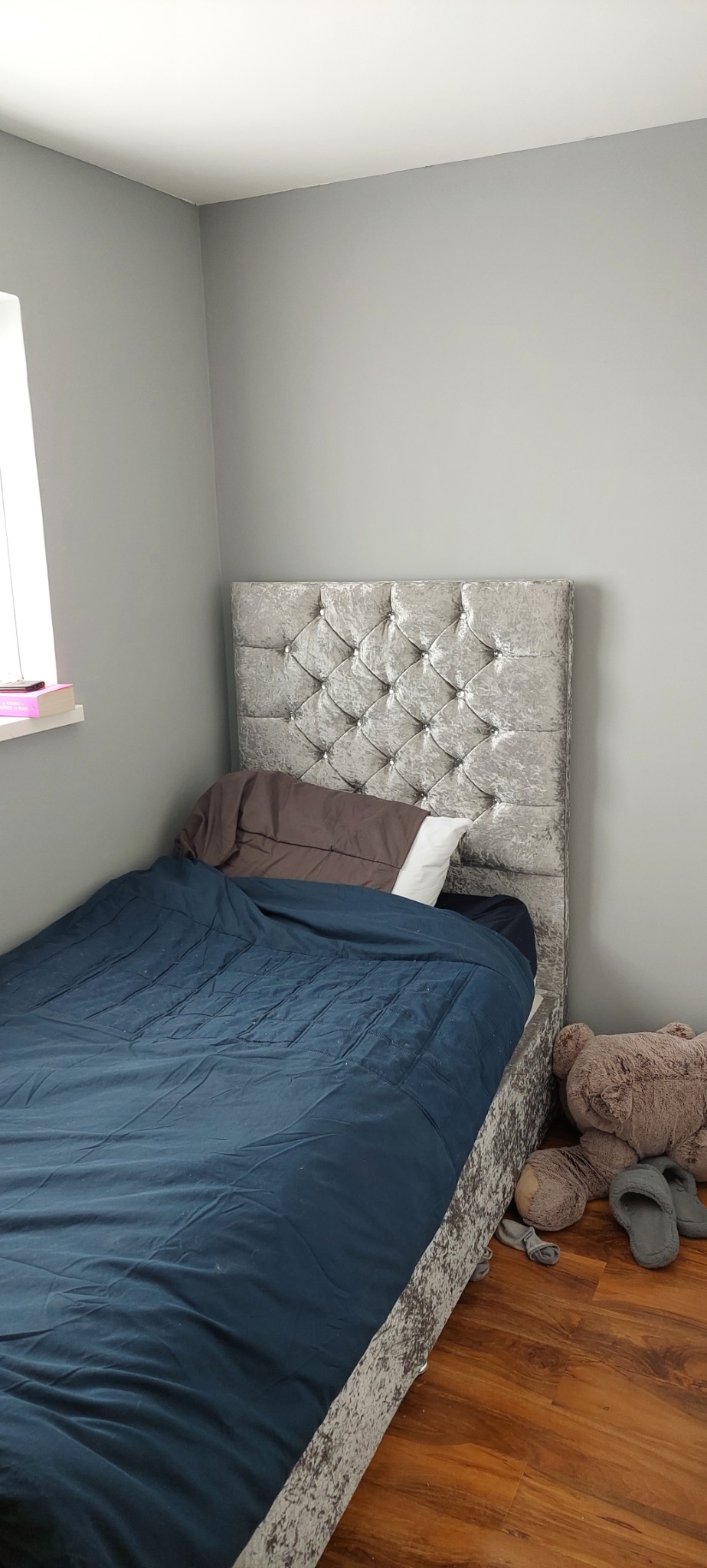 Sypialnia z łóżkiem pojedynczym z srebrnym, pikowanym zagłówkiem, niebieską pościelą i drewnianą podłogą, widoczne pluszowy miś i kapcie.