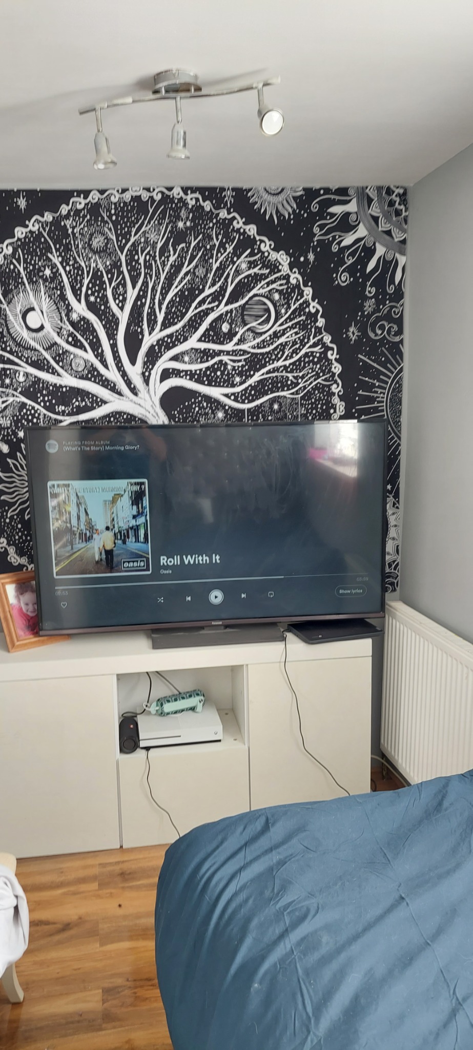 Telewizor z włączoną muzyką Oasis, stojący na białej szafce RTV, nad nim lampa sufitowa i dekoracyjna tapeta z motywem drzewa na czarnym tle.