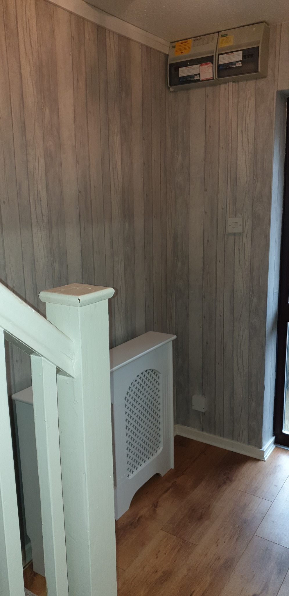 Korytarz z panelami podłogowymi i ścianami wykończonymi tapetą imitującą drewniane deski. Widoczna biała balustrada schodów, obudowa grzejnika i skrzynka elektryczna z naklejkami ostrzegawczymi.