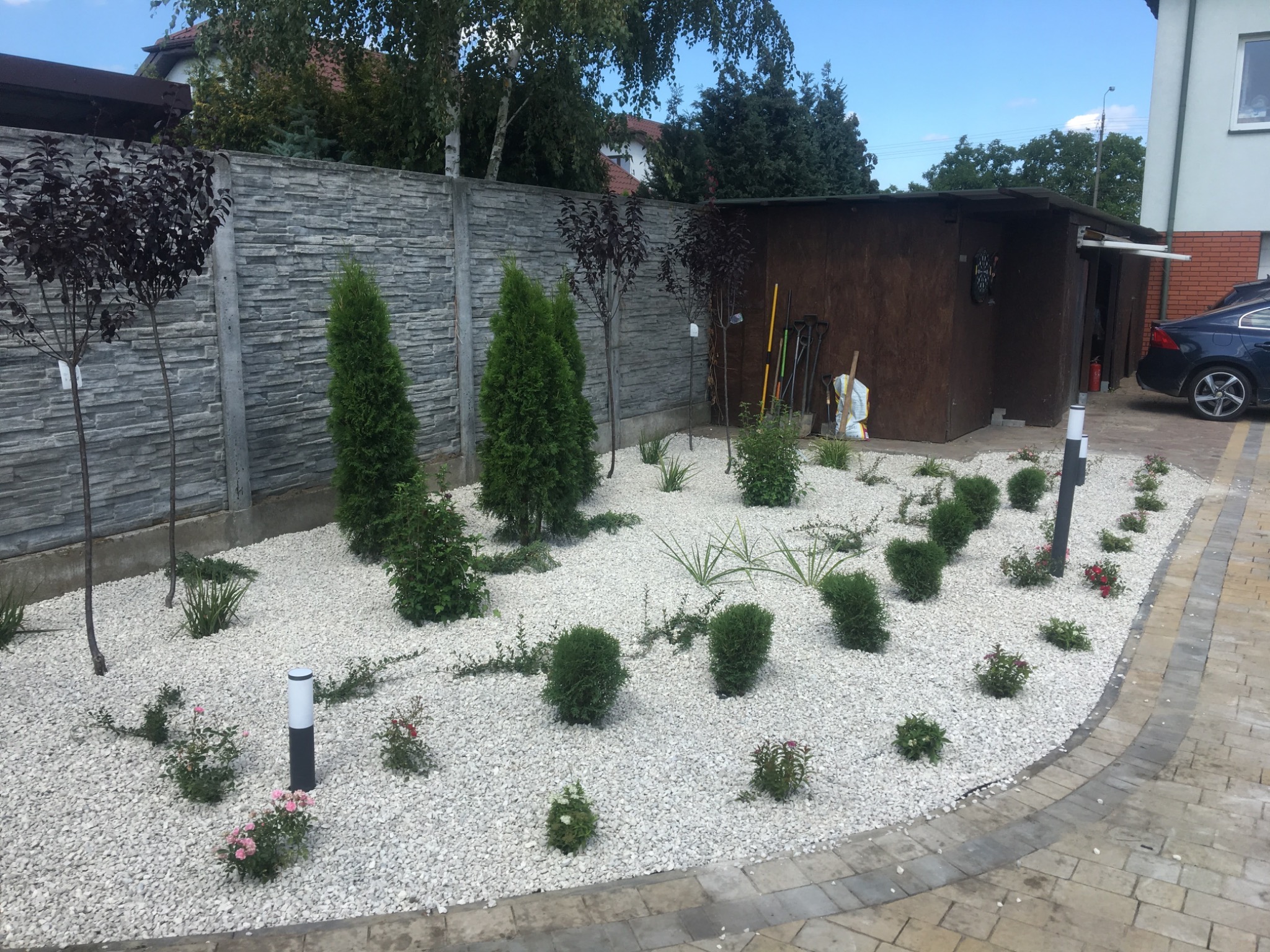 Ogród z białym żwirem, niskimi krzewami i lampami ogrodowymi, obok szopa na narzędzia i zaparkowany samochód.