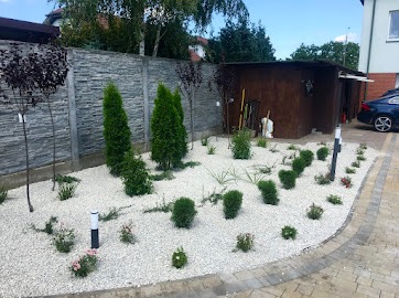 Ogród z białym żwirem, niskimi krzewami i tujami, ogrodzony szarym murem z elementami kamiennymi. Widoczne lampy ogrodowe wzdłuż ścieżki.