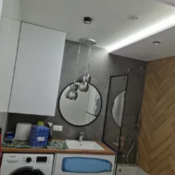 Nowoczesna łazienka z pralką, okrągłym lustrem, wiszącą lampą z trzema chromowanymi kulami i ścianą wyłożoną drewnem w jodełkę, oświetlona listwą LED na suficie.