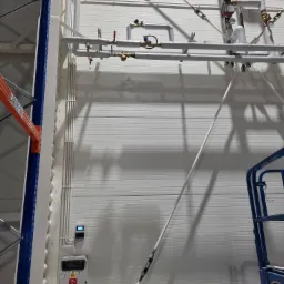 Biała ściana hali z widoczną instalacją rur, zaworów i manometrów, obok pionowy panel elektryczny z gniazdami i wyświetlaczem, fragment konstrukcji regału i podnośnika koszowego.