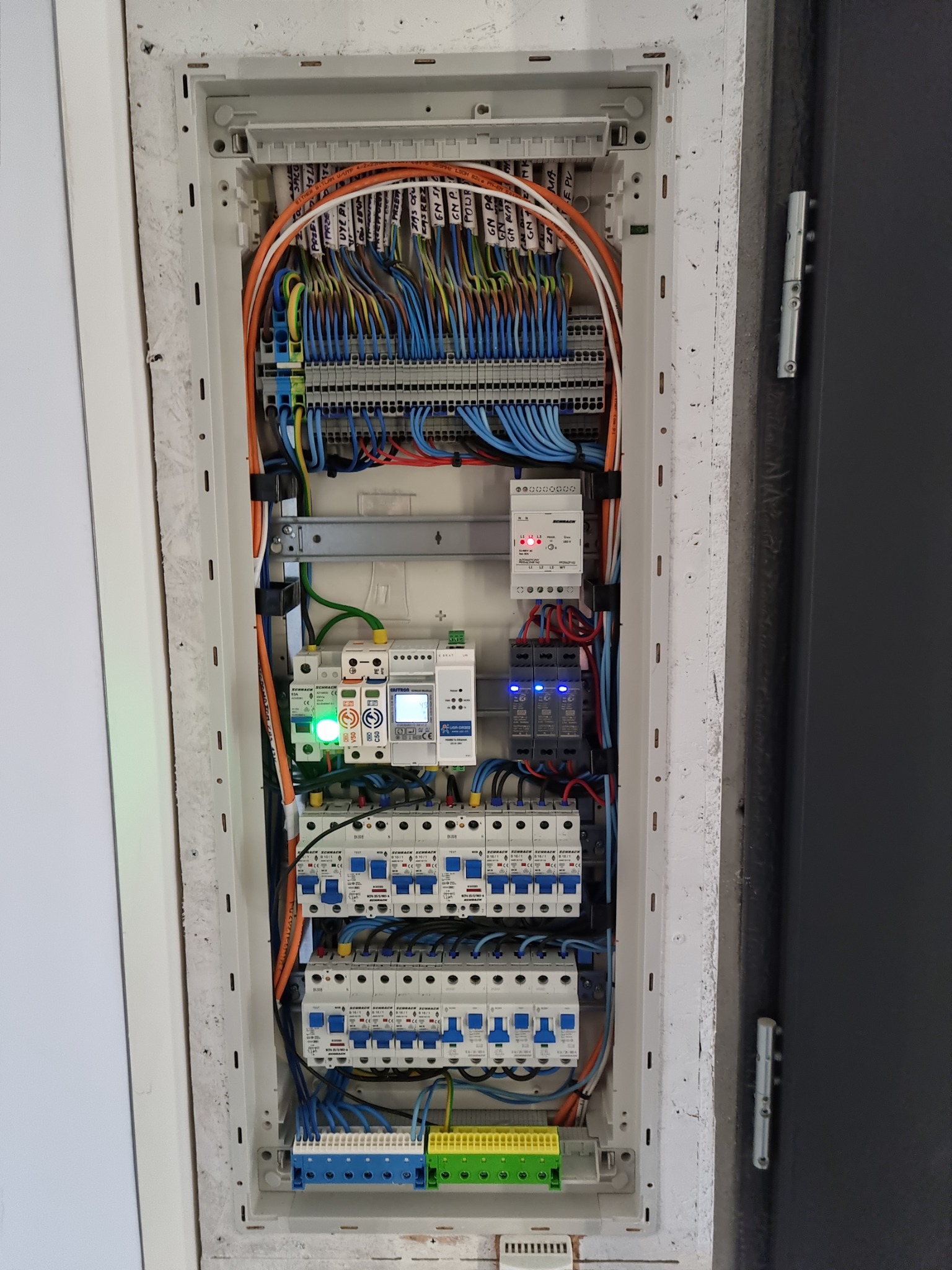 Otwarta skrzynka rozdzielcza z bezpiecznikami, przewodami oznaczonymi etykietami i urządzeniami zabezpieczającymi, zamontowana w ścianie z widoczną strukturą i szarą futryną drzwi po prawej stronie.