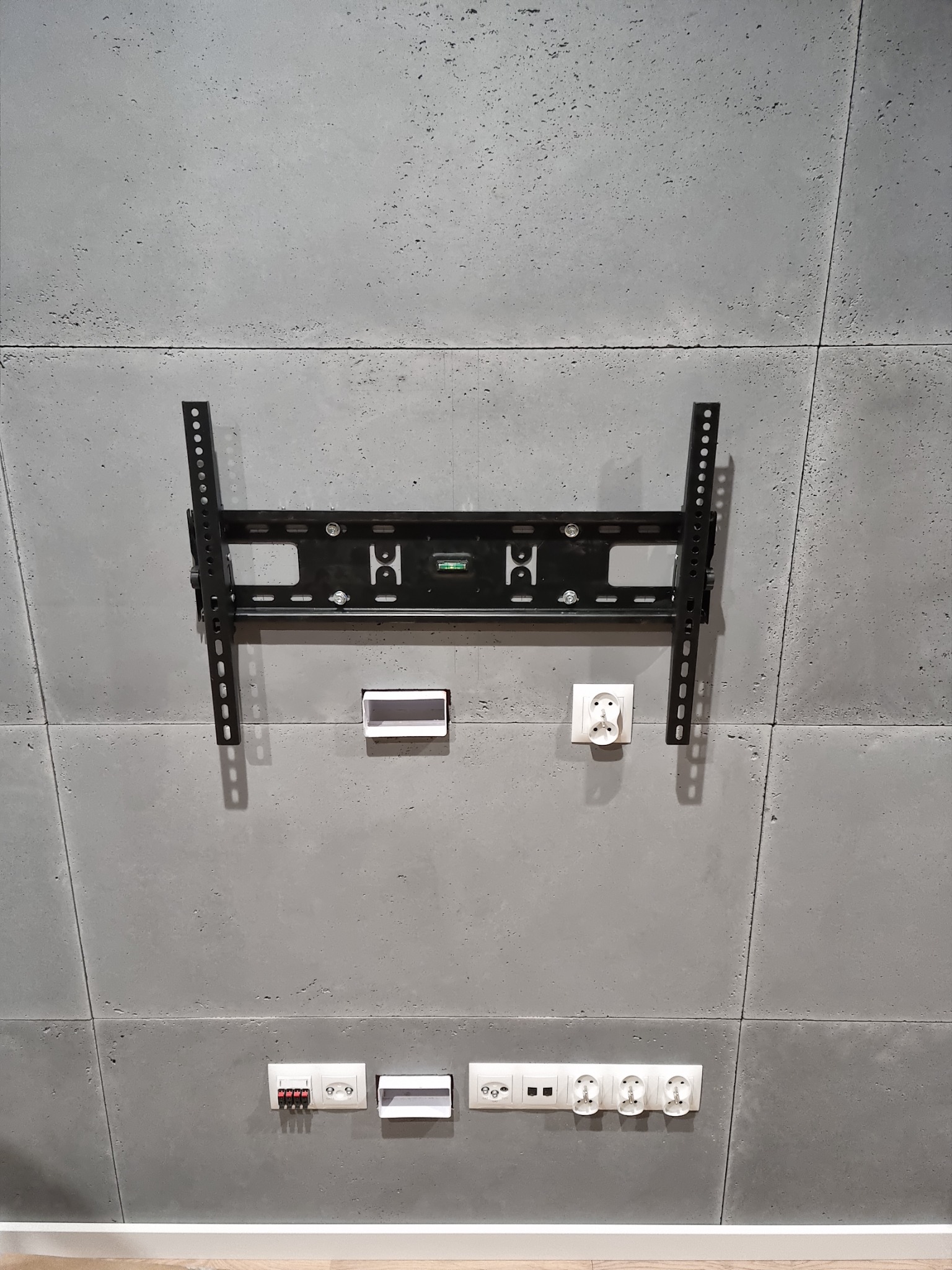 Czarny uchwyt do telewizora zamontowany na szarej, betonowej ścianie w Zabrzu, widoczne gniazdka elektryczne i puszki instalacyjne.