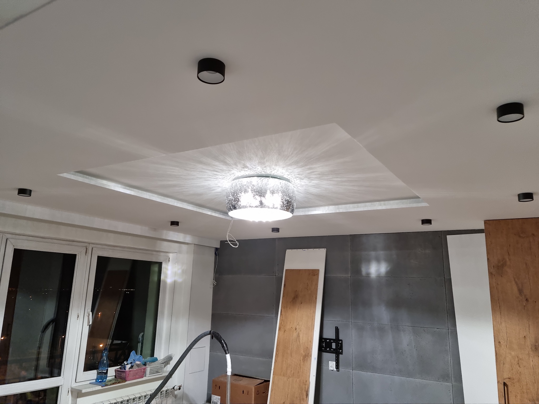 Sufit podwieszany z oświetleniem punktowym i centralnym żyrandolem, widoczne przewody, ściana z szarych paneli, okno z widokiem na miasto nocą, elementy w trakcie montażu.