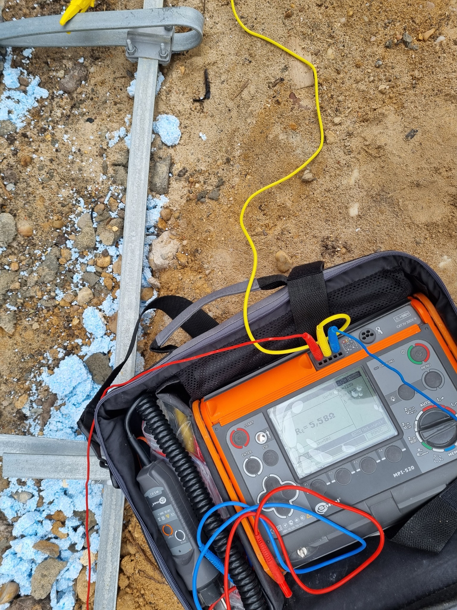 Pomiar rezystancji uziemienia za pomocą miernika Sonel MPI-520 w torbie, widoczne przewody pomiarowe w kolorach żółtym, czerwonym i niebieskim, obok fragment konstrukcji uziomu i rozsypany styropian.