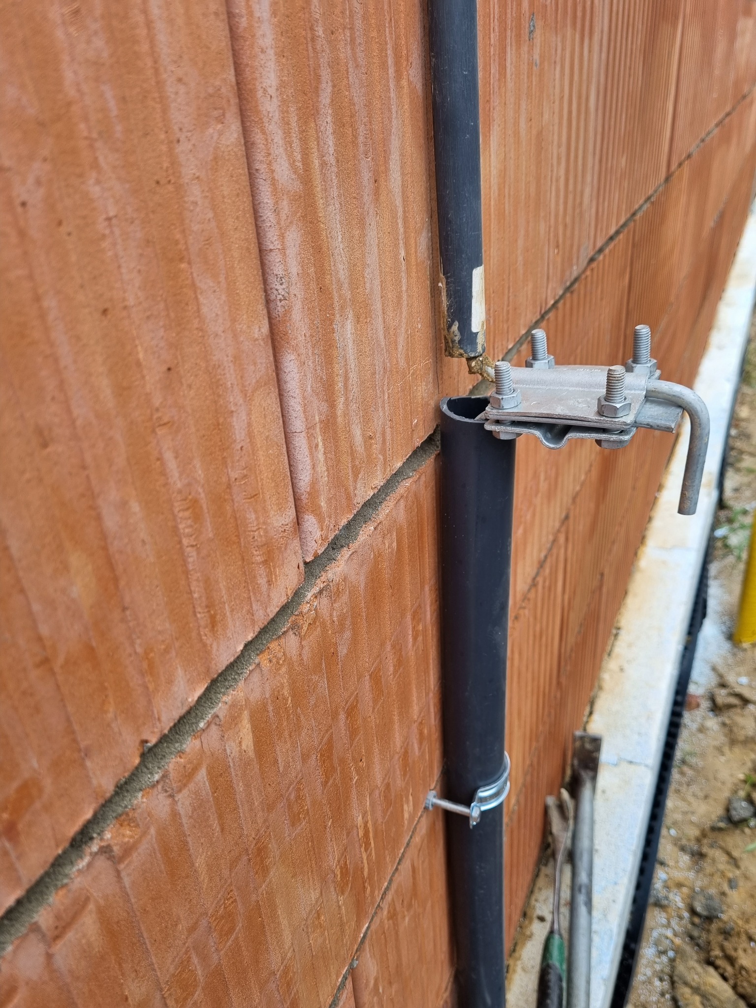 Czarna rura spustowa przymocowana do ściany z czerwonej cegły za pomocą metalowych uchwytów i śrub, widoczne narzędzia przy podstawie ściany.