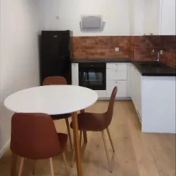 Nowoczesna kuchnia z brązową cegłą na ścianie, białymi szafkami, czarnym blatem i okrągłym białym stołem z brązowymi krzesłami.