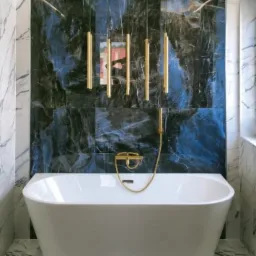 Nowoczesna łazienka z wolnostojącą białą wanną, armaturą w kolorze złotym i płytkami imitującymi marmur na podłodze i ścianach.