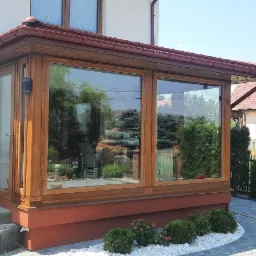 Zadaszone, przeszklone przedłużenie domu z drewnianą konstrukcją, widoczne schody wejściowe z szarej kostki, otoczone białymi kamykami i roślinnością, w tle dom z czerwoną dachówką i zadbany ogród.