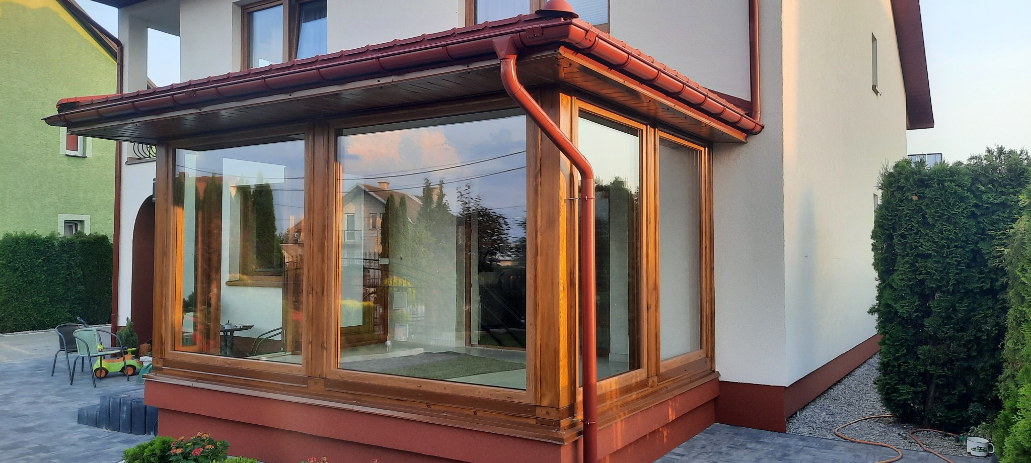 Zadaszony taras z drewnianą konstrukcją i dużymi oknami, z widocznym odbiciem sąsiednich budynków w szybach, czerwona rynna i podmurówka, otoczony zielenią.