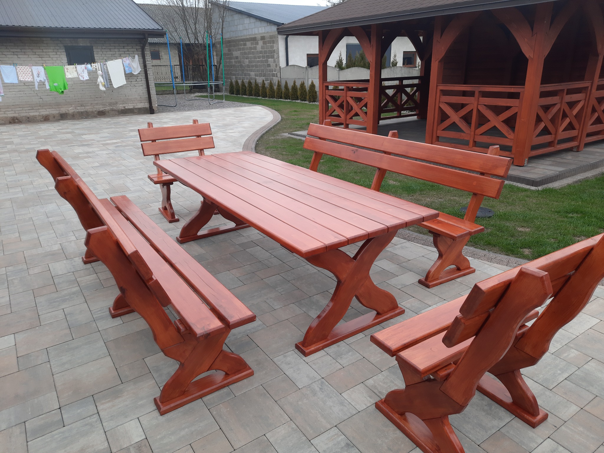 Rustykalny zestaw mebli ogrodowych z drewna: stół i cztery ławki z oparciami, ustawione na brukowanym patio obok drewnianej altany. W tle budynek z suszącym się praniem.