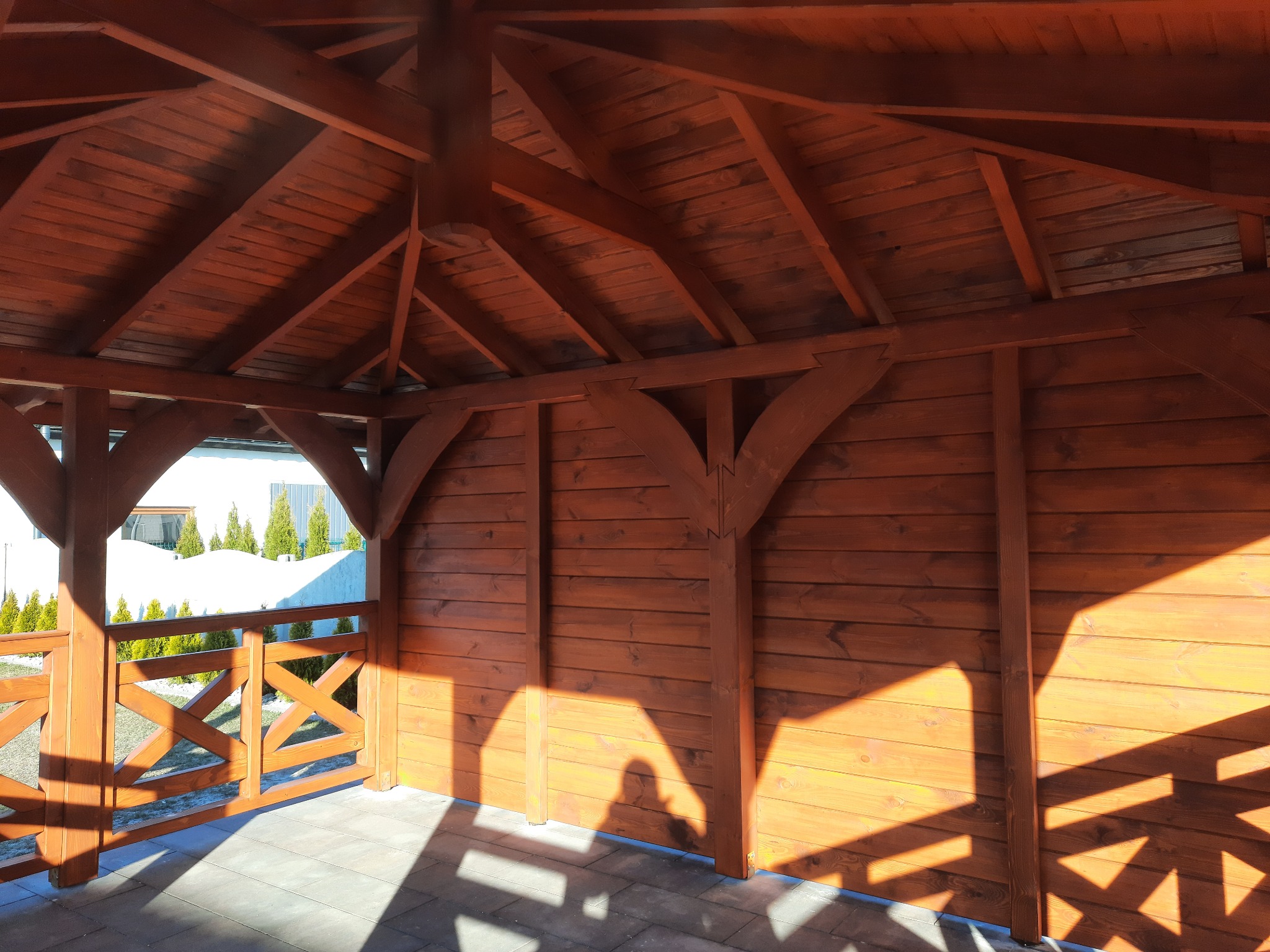 Wnętrze nowej, drewnianej konstrukcji z dachem skośnym, pomalowanej na brązowo, z widocznymi elementami konstrukcyjnymi i zarysami cieni na podłodze z kostki brukowej.