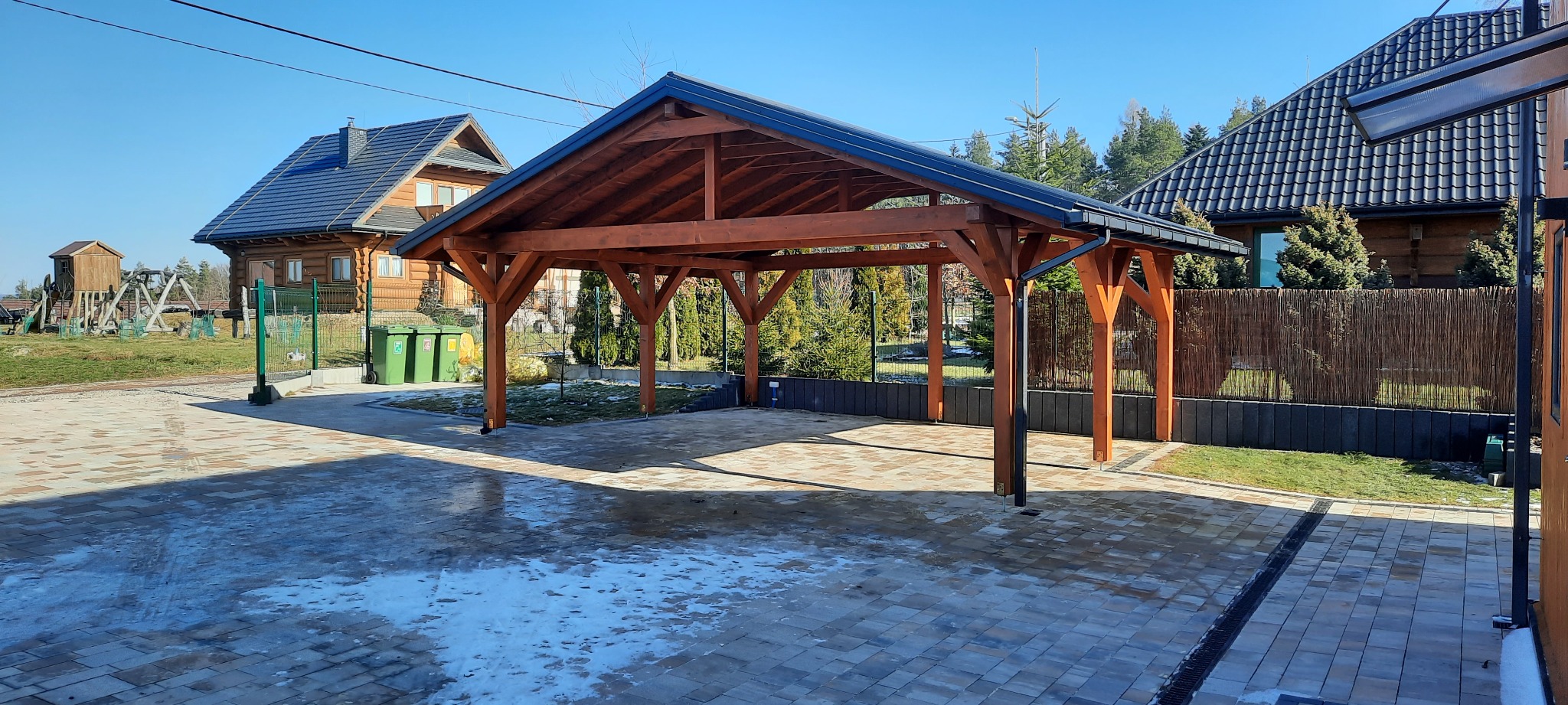 Drewniana wiata garażowa z ciemnym dachem na posesji z kostki brukowej, w tle dom z bali i iglaki.