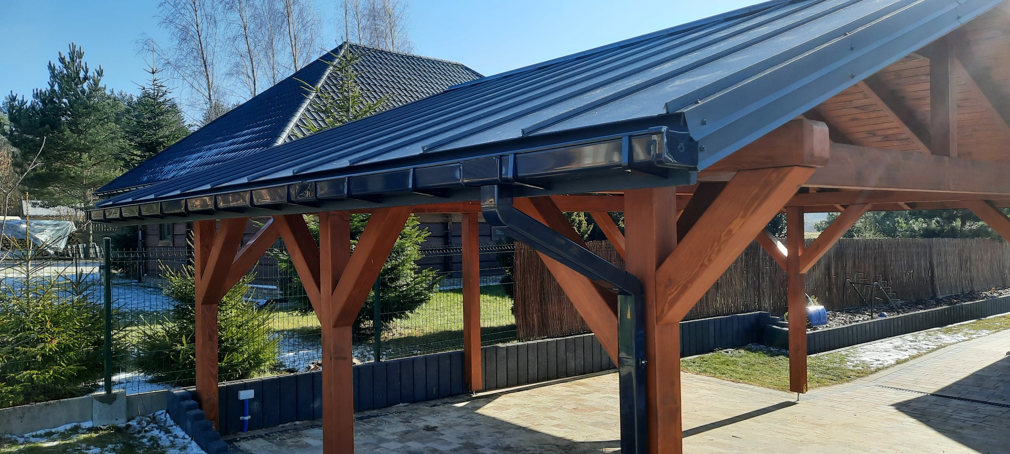 Nowoczesna altana z ciemnym, metalowym dachem i drewnianymi słupami, widok z zewnątrz w słoneczny dzień, na tle domu z ciemnym dachem i zieleni.