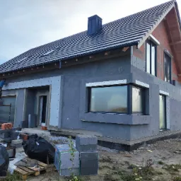 Dom jednorodzinny w trakcie ocieplania elewacji styropianem, widoczne rusztowanie z pracownikiem, czarne worki z odpadami i materiały budowlane na ziemi.