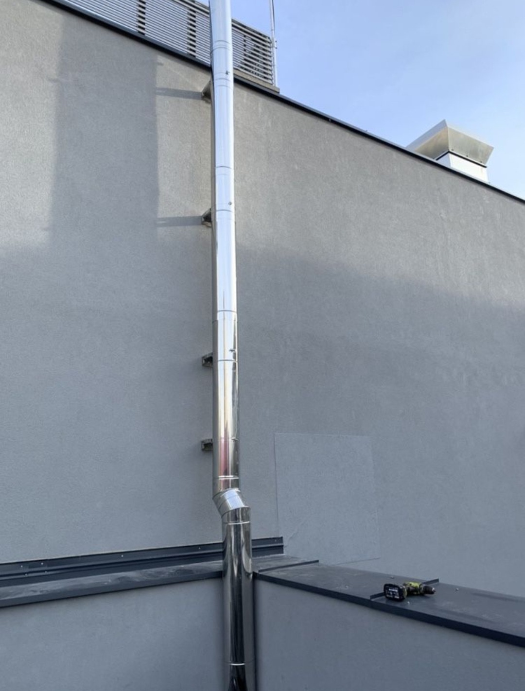 Zewnętrzny, pionowy przewód kominowy ze stali nierdzewnej, zamocowany do szarej ściany budynku, z widocznymi narzędziami na dachu.