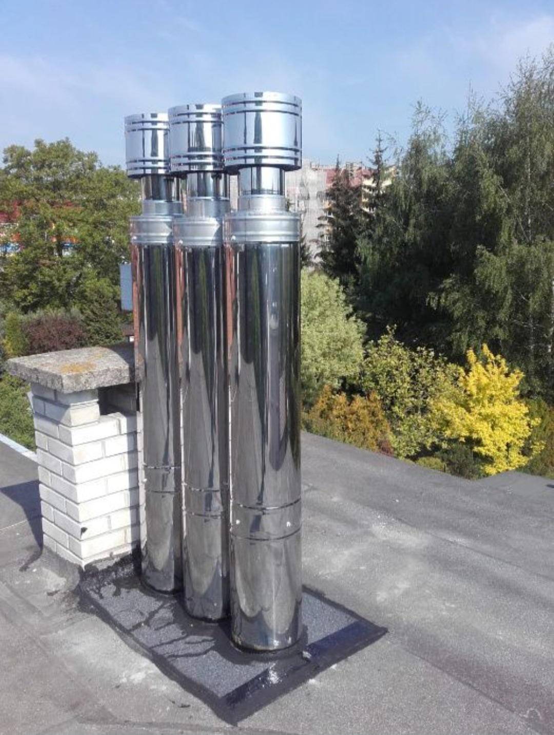 Trzy lśniące, metalowe kominy wentylacyjne zamontowane na płaskim dachu, widoczne elementy uszczelniające i otaczająca zieleń.