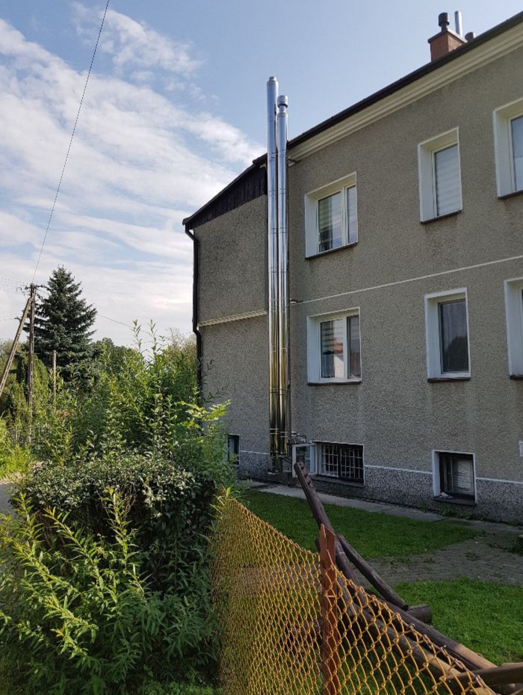 Dwa pionowe, metalowe kominy wentylacyjne przymocowane do bocznej ściany dwupiętrowego budynku mieszkalnego z szarego tynku, widoczne zza żółtego ogrodzenia i zieleni.