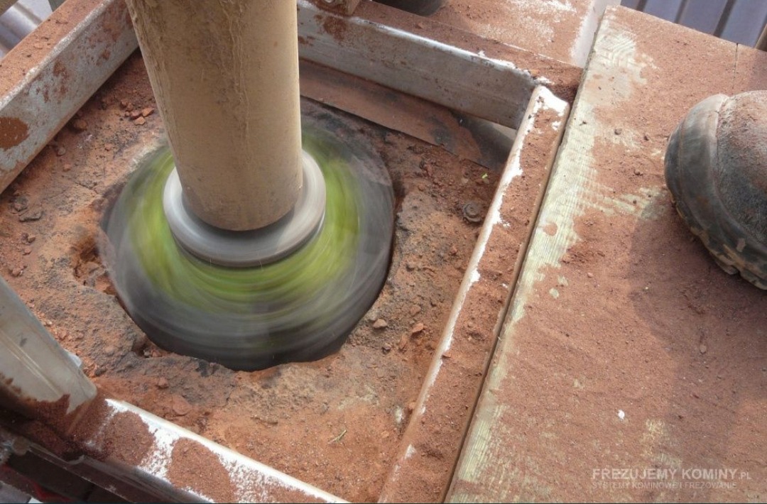 Frezowanie komina: obracająca się głowica frezarska z zielonymi elementami tnącymi w trakcie pracy, widoczne pył i urobek, fragment buta roboczego w prawym górnym rogu.