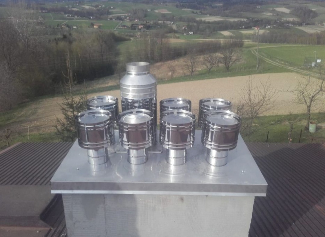 Komplet nowych, błyszczących, stalowych nasad kominowych różnej wielkości, zamontowanych na szczycie komina na tle wiejskiego krajobrazu z polami i drzewami.