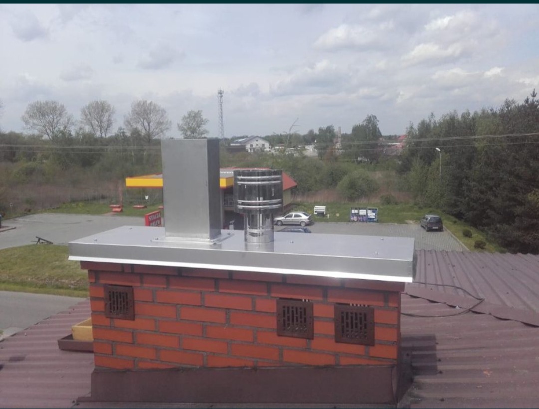 Murowany komin z metalowym wkładem i nasadą wentylacyjną na dachu budynku, widok z góry na otoczenie z drzewami i budynkami.