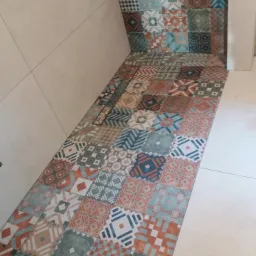 Świeżo ułożona, wielobarwna mozaika z kwadratowych płytek na podłodze i ścianie wnęki łazienki, kontrastująca z jasnymi, gładkimi płytkami na pozostałej powierzchni ścian.