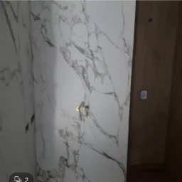 Ściana łazienki wyłożona białymi płytkami imitującymi marmur z widocznymi złotymi bateriami i fragmentem drewnianej ściany z gniazdkiem.