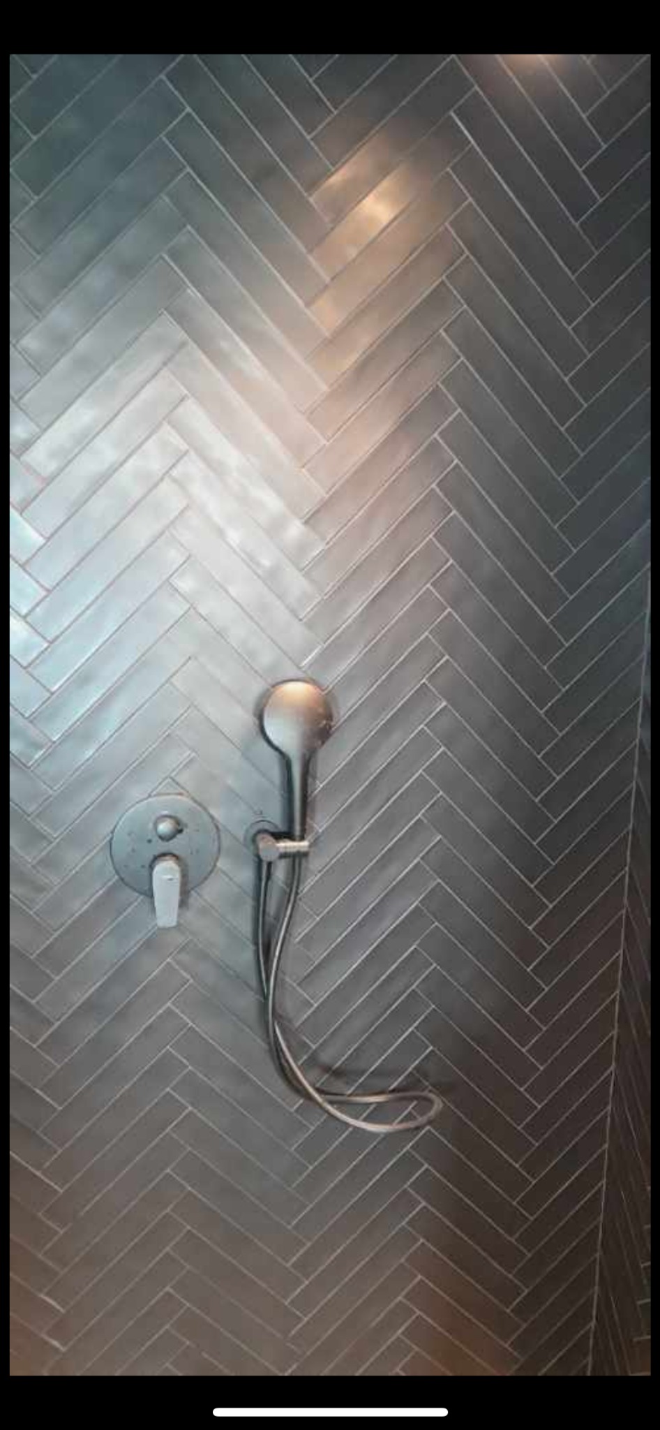 Szara ściana prysznicowa wyłożona płytkami w jodełkę, z widoczną baterią i słuchawką prysznicową. Minimalistyczny design, nowoczesne wykończenie łazienki.