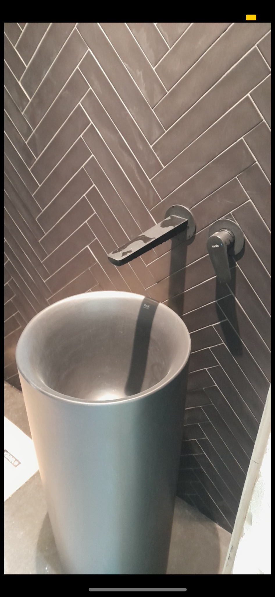 Nowoczesna umywalka wolnostojąca w szarym odcieniu, z czarną armaturą na tle ściany wyłożonej ciemnymi, wąskimi płytkami w jodełkę. Minimalistyczny design.