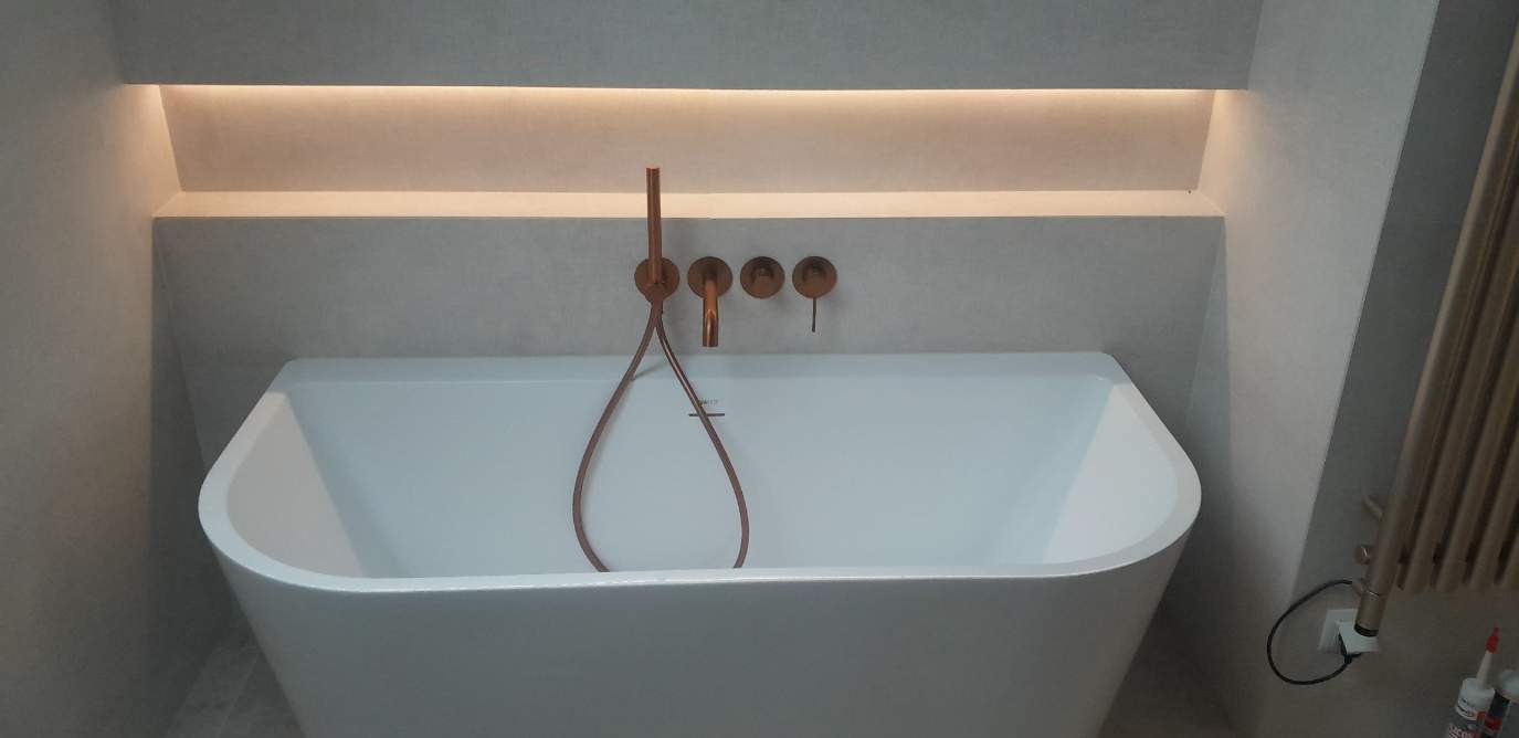 Nowoczesna łazienka: biała wanna wolnostojąca z miedzianą armaturą na tle szarej ściany z podświetlaną wnęką. Minimalistyczny design, ciepłe oświetlenie.