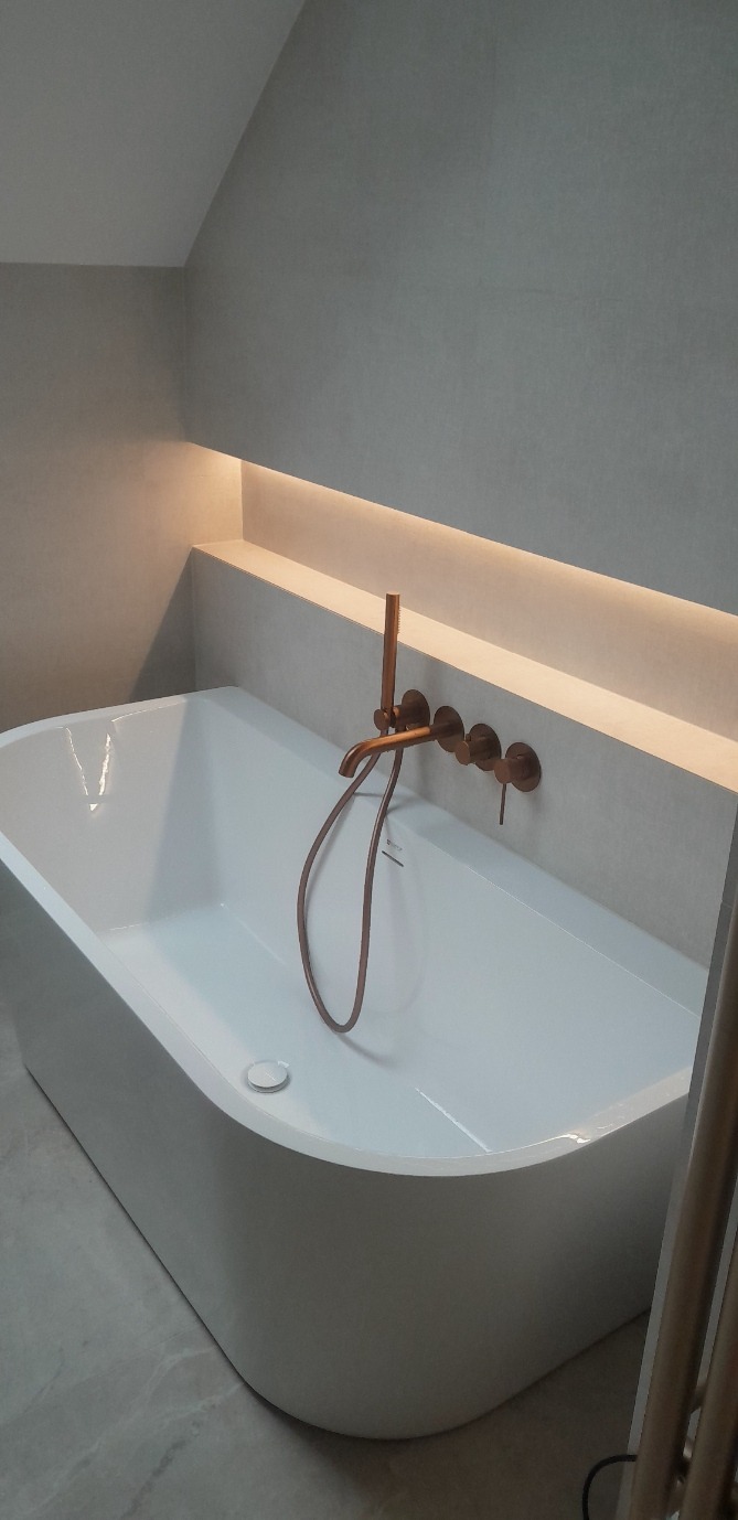 Nowoczesna, biała wanna wolnostojąca z miedzianą armaturą na tle szarej ściany z podświetleniem LED. Minimalistyczny design łazienki na poddaszu.