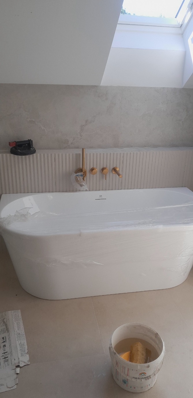 Nowa, biała wanna owinięta folią ochronną w łazience z beżowymi płytkami i złotą armaturą. Widoczny fragment okna dachowego i narzędzia glazurnicze.