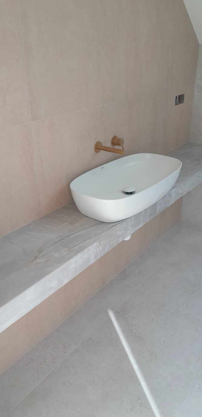 Nowoczesna łazienka z owalną umywalką nablatową i złotą armaturą na tle beżowych płytek. Minimalistyczny design, jasne wnętrze.