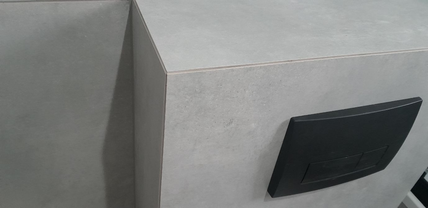 Szara zabudowa WC z czarnym przyciskiem spłukującym, wykończona płytkami o jednolitej fakturze. Minimalistyczny design, precyzyjne łączenia płytek.