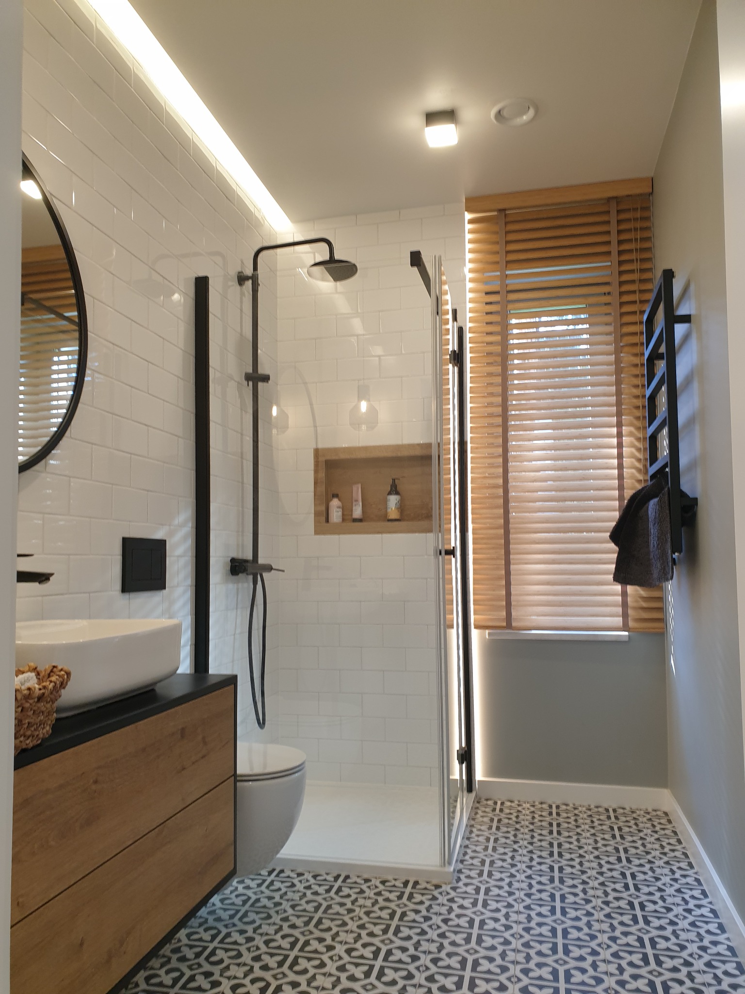 Nowoczesna łazienka z białymi kaflami, czarną armaturą, drewnianymi żaluzjami i wzorzystą podłogą, oświetlona listwą LED w suficie.