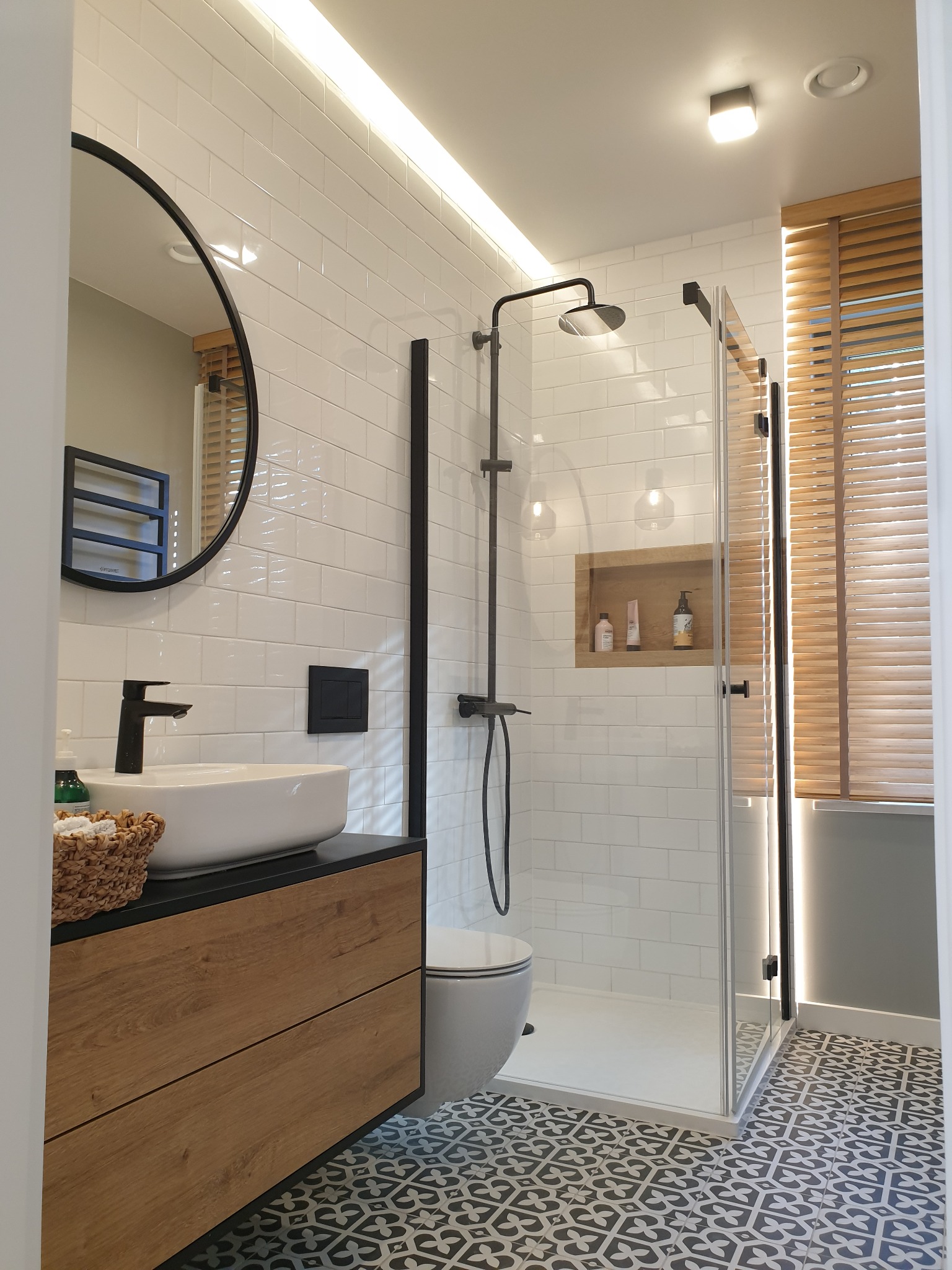 Nowoczesna łazienka z kabiną prysznicową typu walk-in, umywalką nablatową na drewnianej szafce z czarnym blatem, okrągłym lustrem i wzorzystą podłogą w odcieniach szarości.