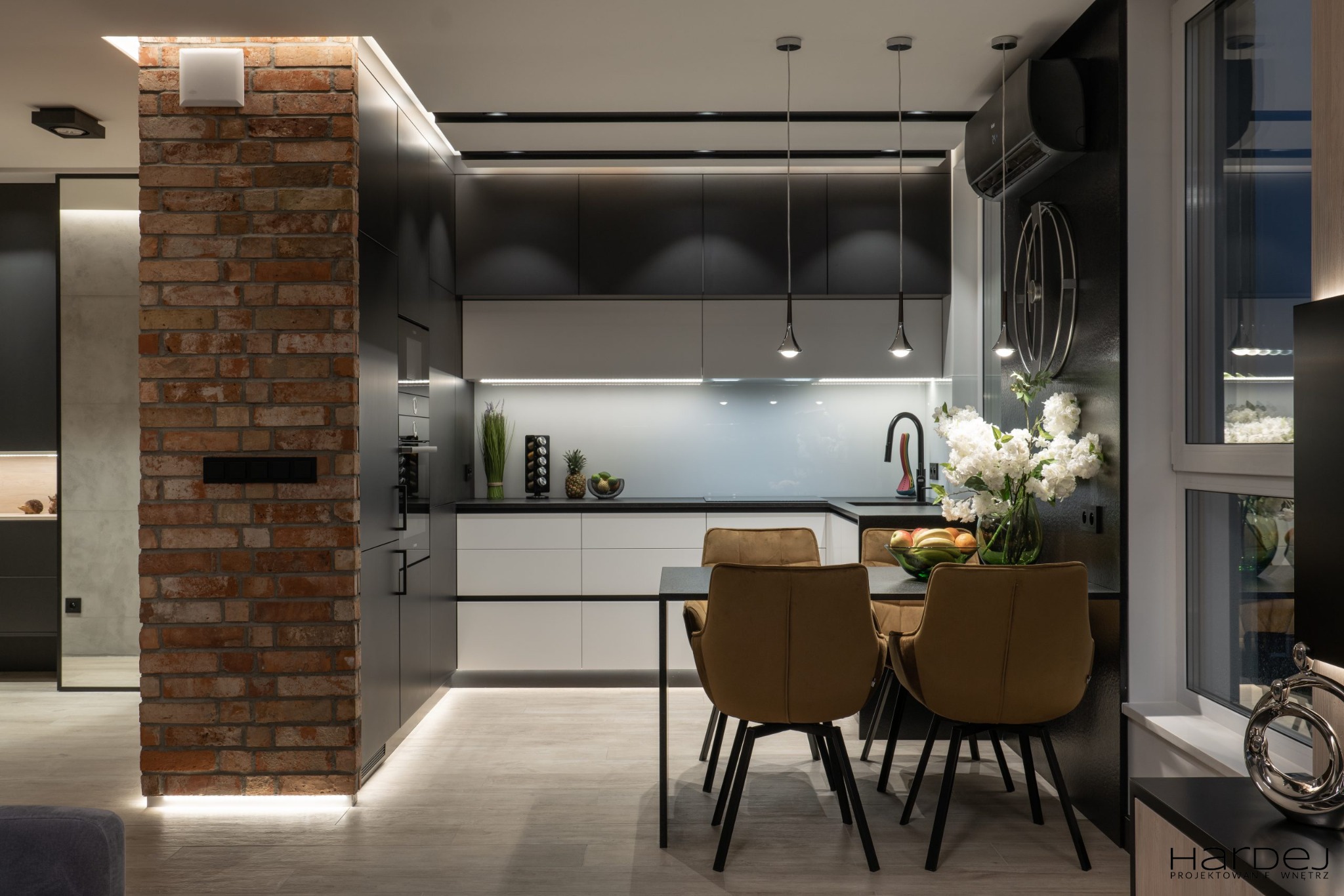 Nowoczesna kuchnia z ceglanym filarem, czarno-białymi szafkami, oświetleniem LED pod szafkami i wiszącymi lampami nad stołem jadalnym z krzesłami w kolorze musztardowym.
