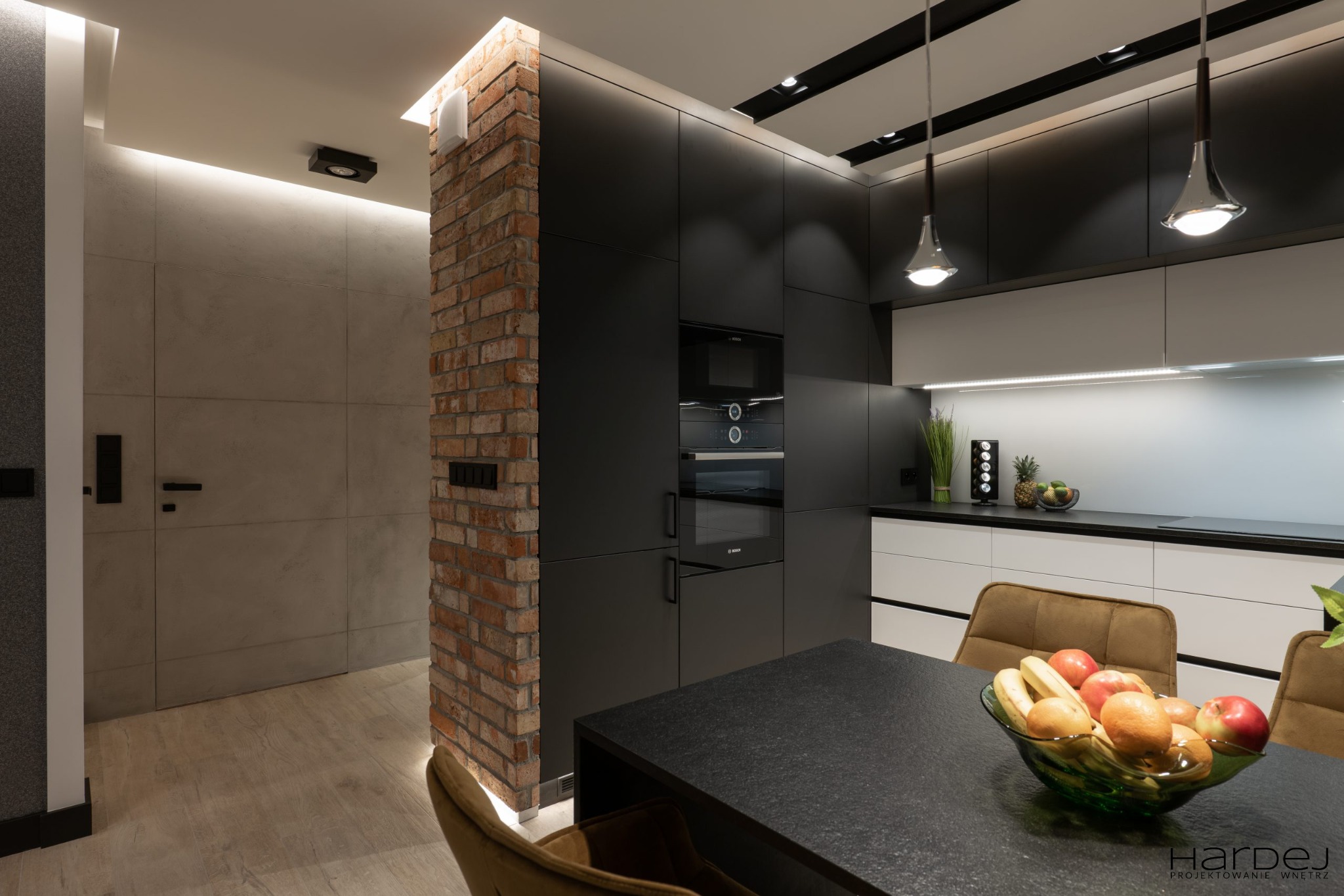 Nowoczesna kuchnia z czarnymi szafkami, ceglanym filarem i czarnym blatem stołu, oświetlona lampami wiszącymi i ukrytym światłem LED.