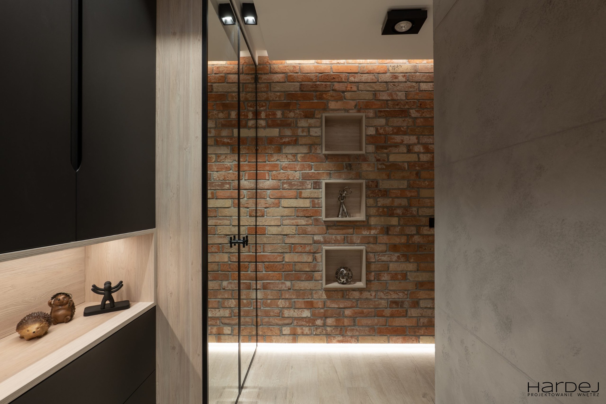 Nowoczesne wnętrze z ceglaną ścianą, podświetlanymi półkami z dekoracjami, czarnymi szafkami i drewnianymi akcentami, widziane z perspektywy korytarza.