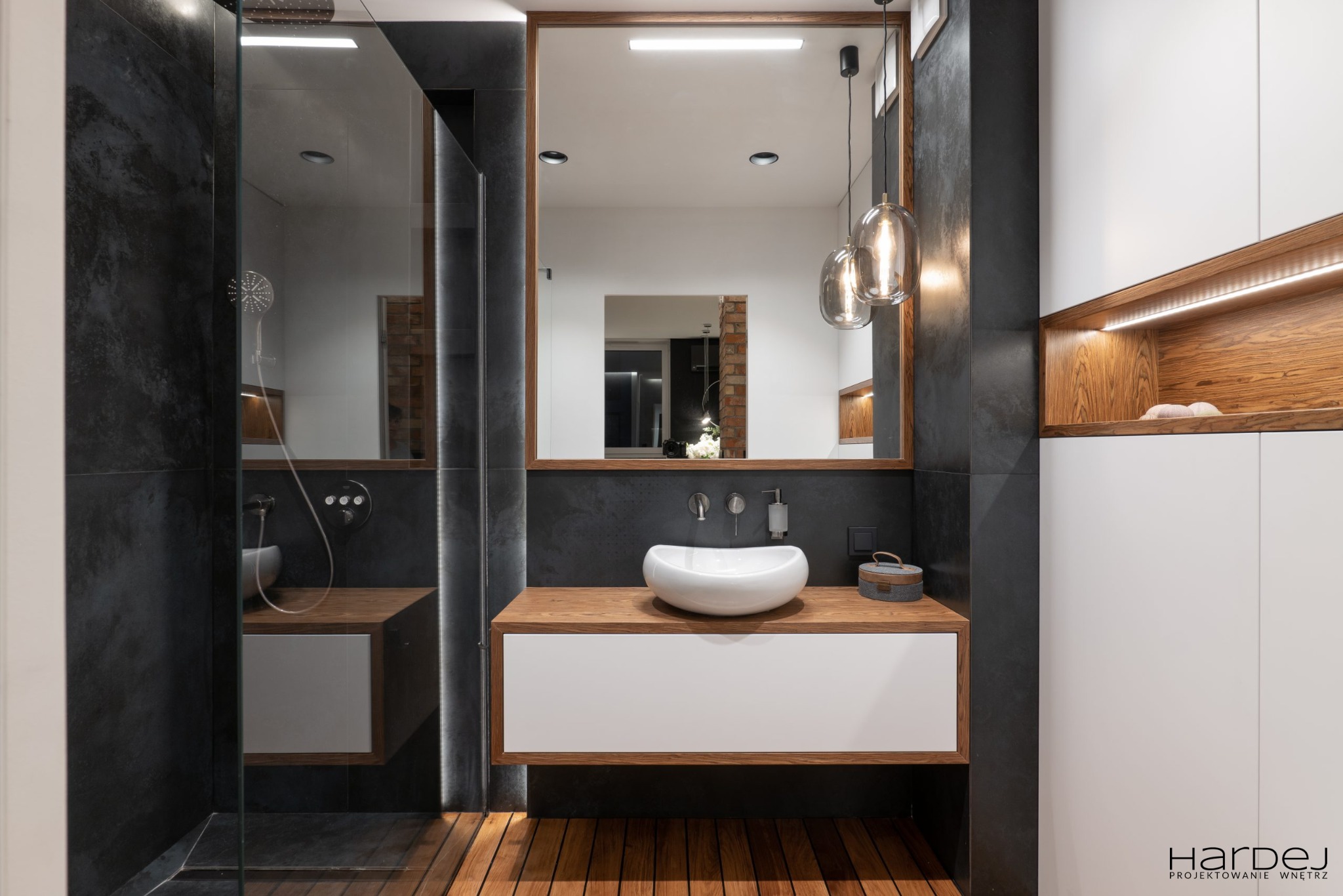 Nowoczesna łazienka z drewnianą podłogą, umywalką nablatową na blacie z drewna, lustrem w drewnianej ramie i czarnymi płytkami na ścianach, oświetlenie LED.