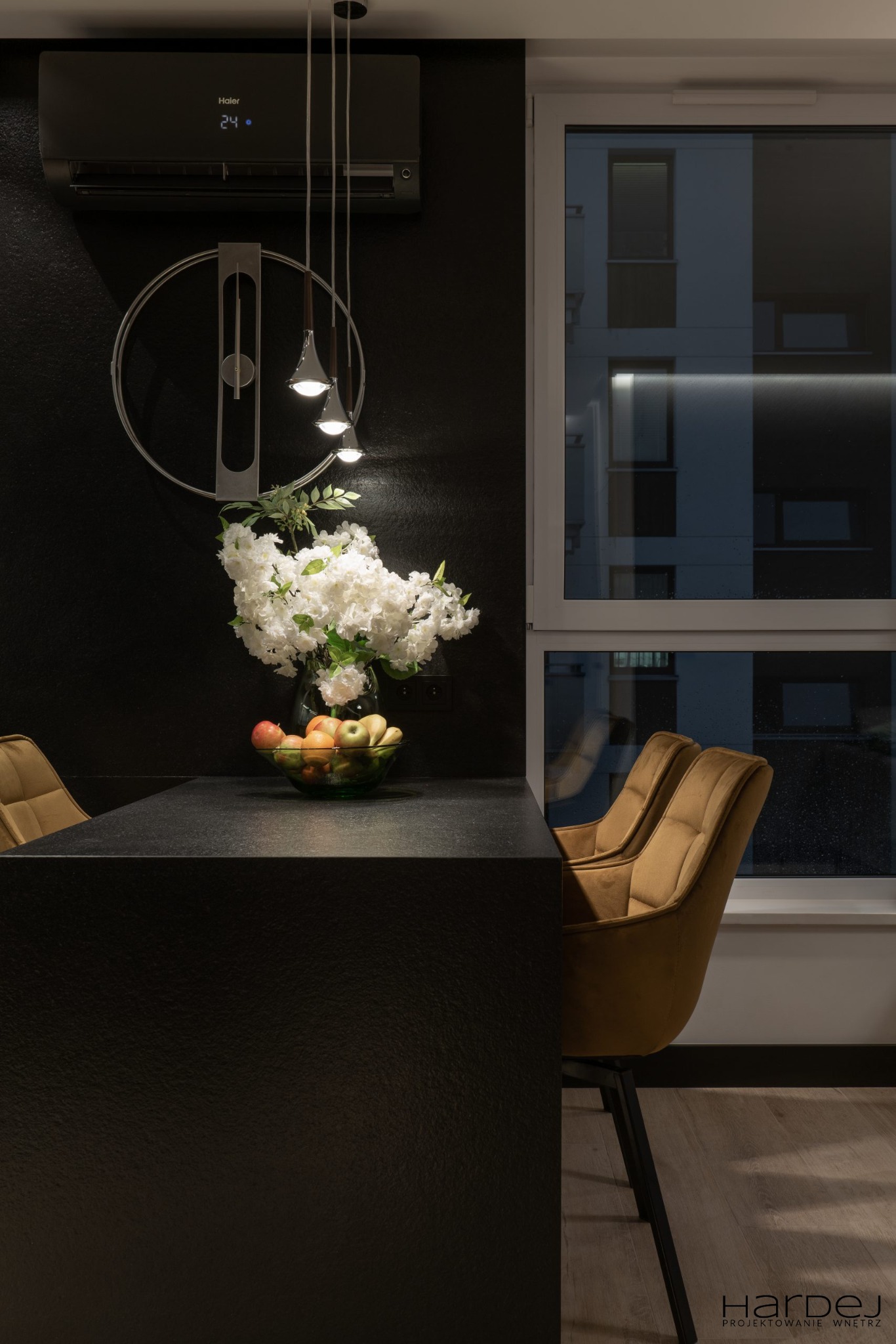 Ciemna kuchnia z czarnym blatem, owocami w szklanej misie, kwiatami i złotymi krzesłami na tle okna z widokiem na budynek.