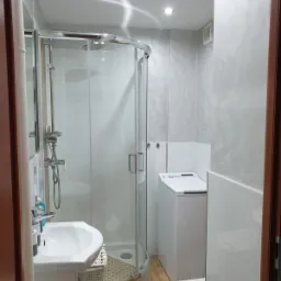 Funkcjonalna łazienka po remoncie: kabina prysznicowa z chromowanymi elementami, umywalka z szafką z wikliny, pralka i toaleta w jasnych barwach, ściany wykończone dekoracyjnym tynkiem.